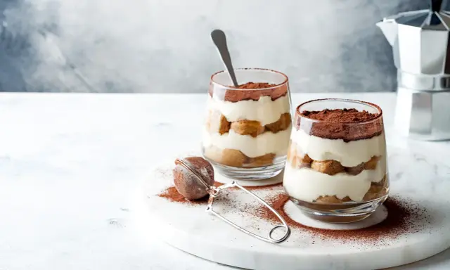 Dwa pucharki z tiramisu bez jajek, posypane kakao, na marmurowej desce.