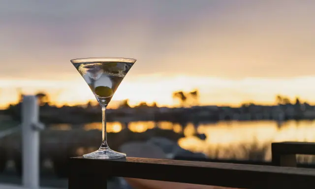 Idealny martini wstrząśnięte nie mieszane, z oliwką, na tle zachodzącego słońca nad wodą.