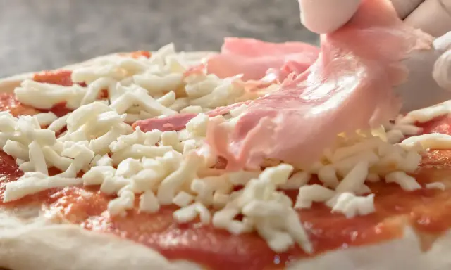 Ręce w białych rękawiczkach układają cienkie plastry szynki do pizzy na serze i sosie pomidorowym.