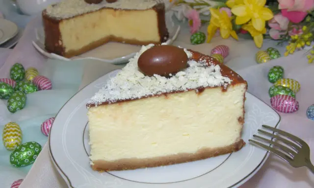Pyszny sernik z mascarpone, udekorowany czekoladowymi jajeczkami i wiórkami białej czekolady, na tle wielkanocnych ozdób.