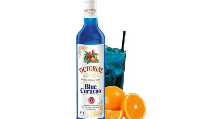 Syrop Blue Curacao Victoria's, idealny do drinków. W szklance niebieski koktajl z lodem i słomką, obok pomarańcze.