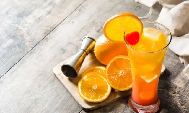 Kolorowy drink tequila sunrise z pomarańczą i wisienką, obok pomarańcze i miarka barmańska.
