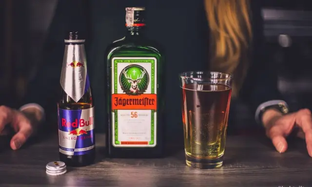 Jägermeister z czym pić? Klasycznie z Red Bullem, tworząc popularny "Jägerbomb". Na stole butelka Jägermeistera, Red Bull i szklanka z drinkiem.