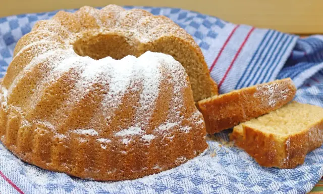 Puszysta babka wielkanocna, oprószona cukrem pudrem, leży na niebiesko-białym ręczniku. Obok niej kilka kromek ciasta.
