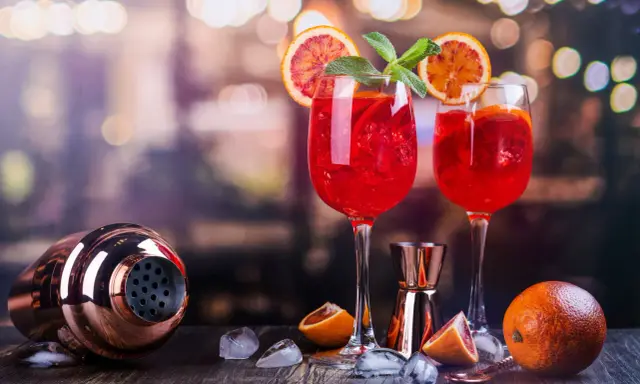 Dwa kieliszki orzeźwiającego drinka z prosecco z czymś czerwonym, plasterkami pomarańczy i miętą. Obok shaker i pomarańcze.