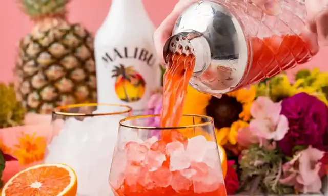 Malibu z sokiem ananasowym nalewany do szklanki z lodem. W tle ananas, butelka Malibu i kwiaty.