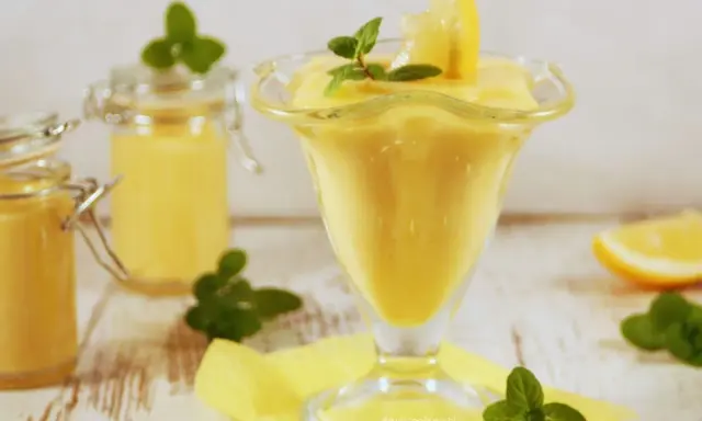 Aksamitny lemon curd w pucharku, ozdobiony plasterkiem cytryny i miętą. Obok słoiczki z tym samym kremowym przysmakiem.