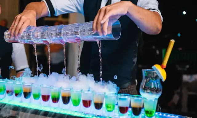 Barman nalewa kolorowe shoty z wielopoziomowego shakera. Efektowny drink bar diy z dymem i neonowym oświetleniem.