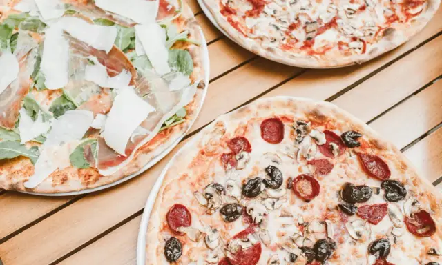 Trzy pizze na drewnianym stole. Jedna z szynką parmeńską i rukolą, druga z salami i pieczarkami, trzecia z pieczarkami. Szukasz alternatywy, jaki ser do pizzy zamiast mozzarelli?