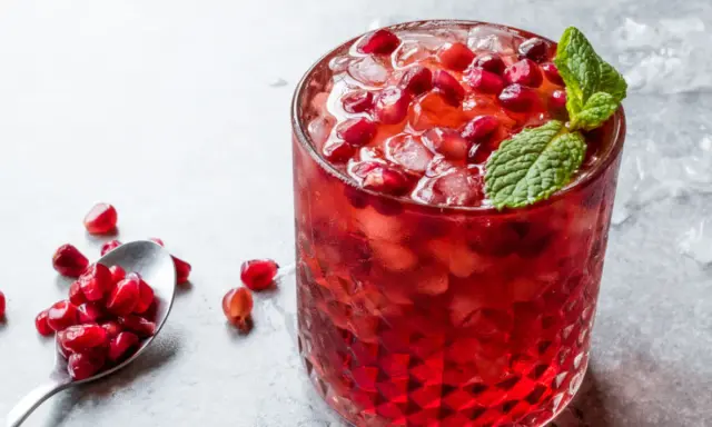 Orzeźwiający drink z grenadyną, lodem i pestkami granatu, ozdobiony listkiem mięty. Obok łyżeczka z owocem.
