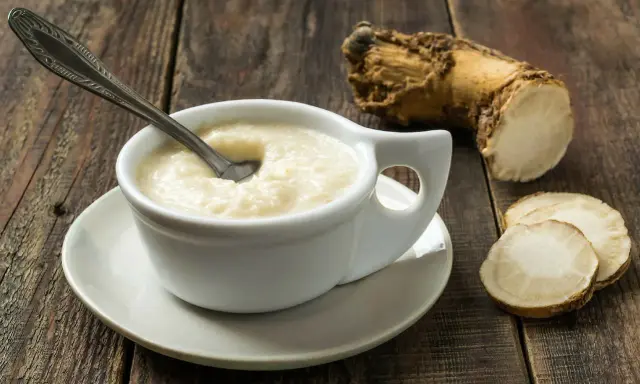 Biały sos chrzanowy w naczyniu, obok korzeń i plasterki chrzanu na drewnianym stole.