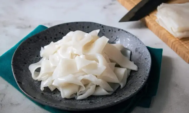 Gotowy do przygotowania makaron ryżowy na talerzu. Obok deska z nożem i stos świeżego makaronu.