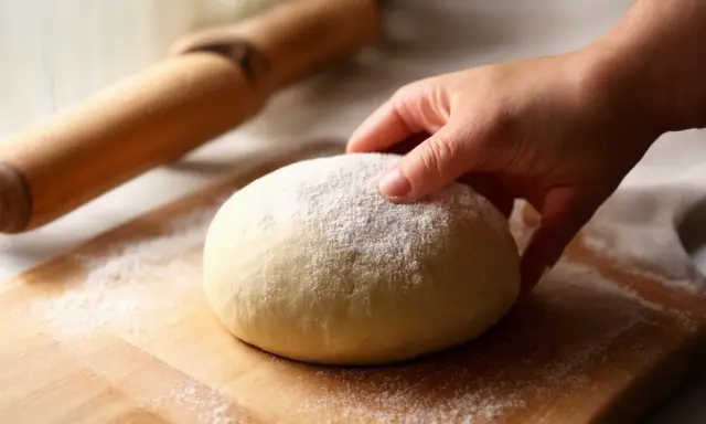 Dłoń dotyka kulki ciasta na pizzę, posypanej mąką. Obok wałek. Czy ciasto na pizzę można mrozić? Tak, można je zamrozić.