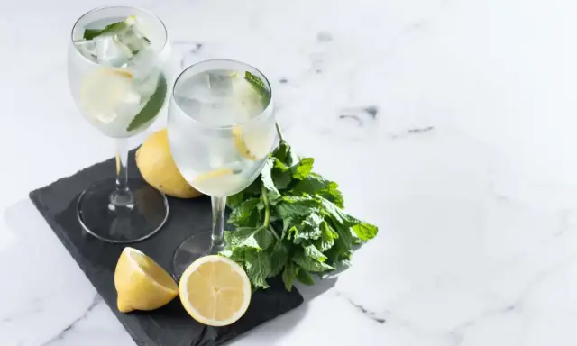 Dwa orzeźwiające drinki z prosecco, lodem, cytryną i miętą, prezentowane na czarnej desce z dodatkiem świeżych cytryn i gałązki mięty.