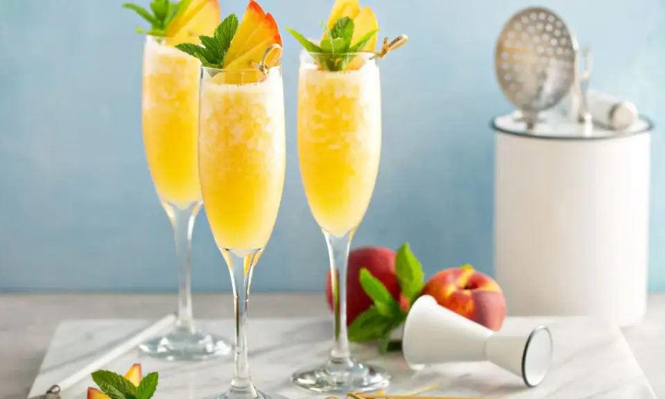 Trzy orzeźwiające drinki mimoza w kieliszkach flute, ozdobione brzoskwinią i miętą, na marmurowym blacie.
