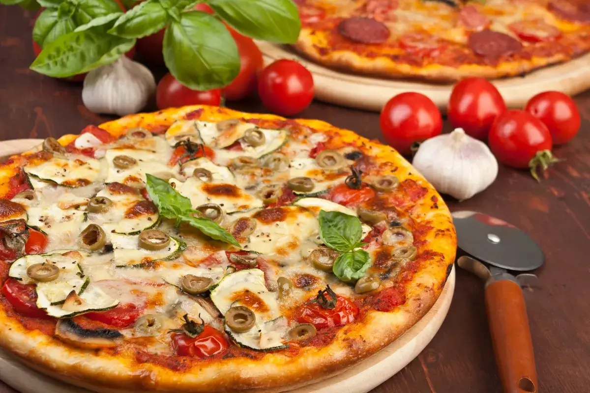 Pizza z cukinią, oliwkami i pomidorkami, obok surowe pieczarki na pizze, czosnek i bazylia.