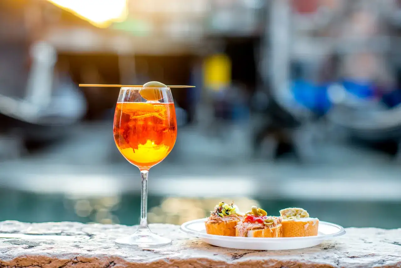 Orzeźwiający drink z Campari i Aperol, ozdobiony oliwką, na tle kanału. Obok talerzyk z pysznymi bruschettami.