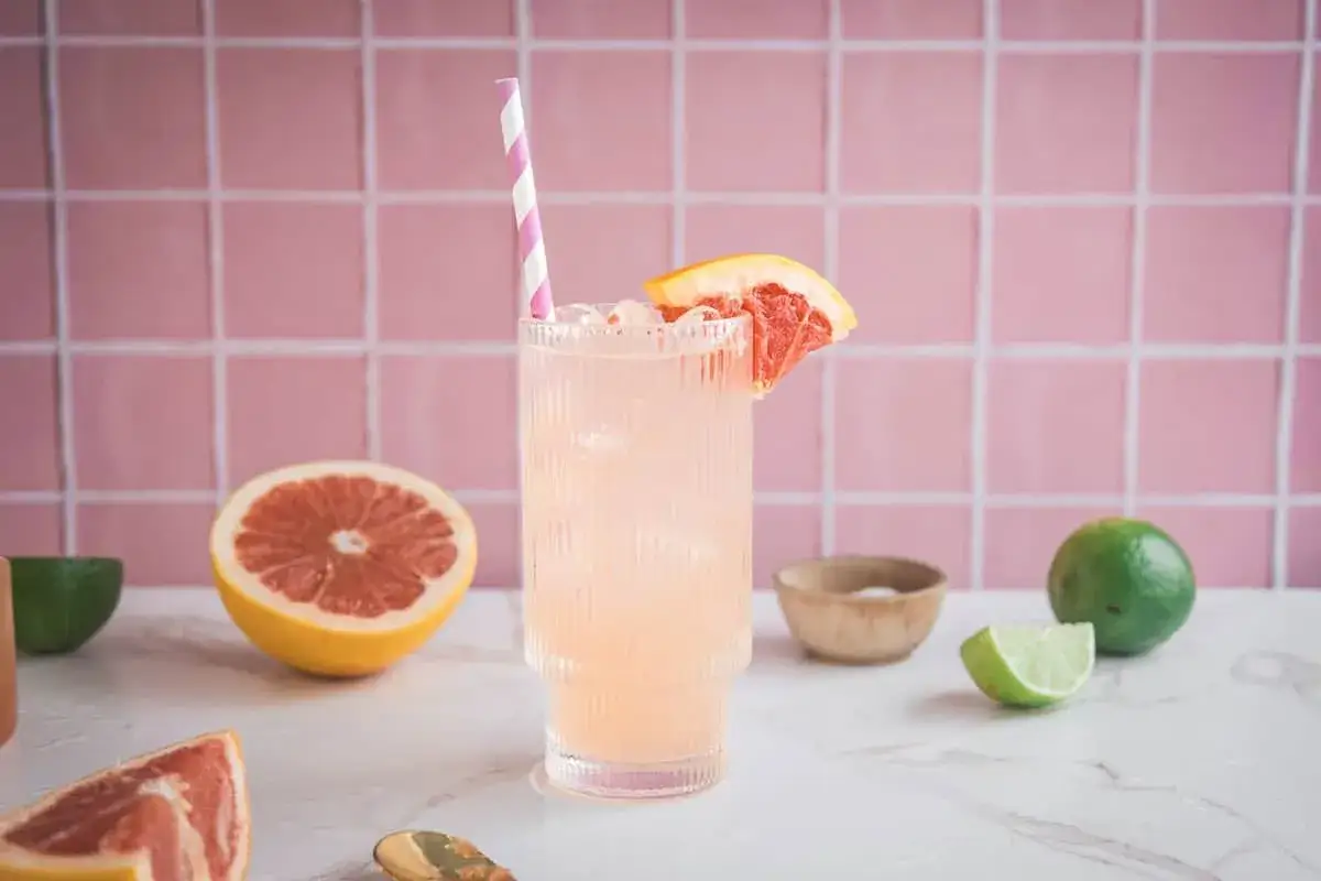 Orzeźwiający drink z grejpfrutem, lodem i plasterkiem owocu, ozdobiony różowo-białą słomką. W tle różowe kafelki i świeże owoce.