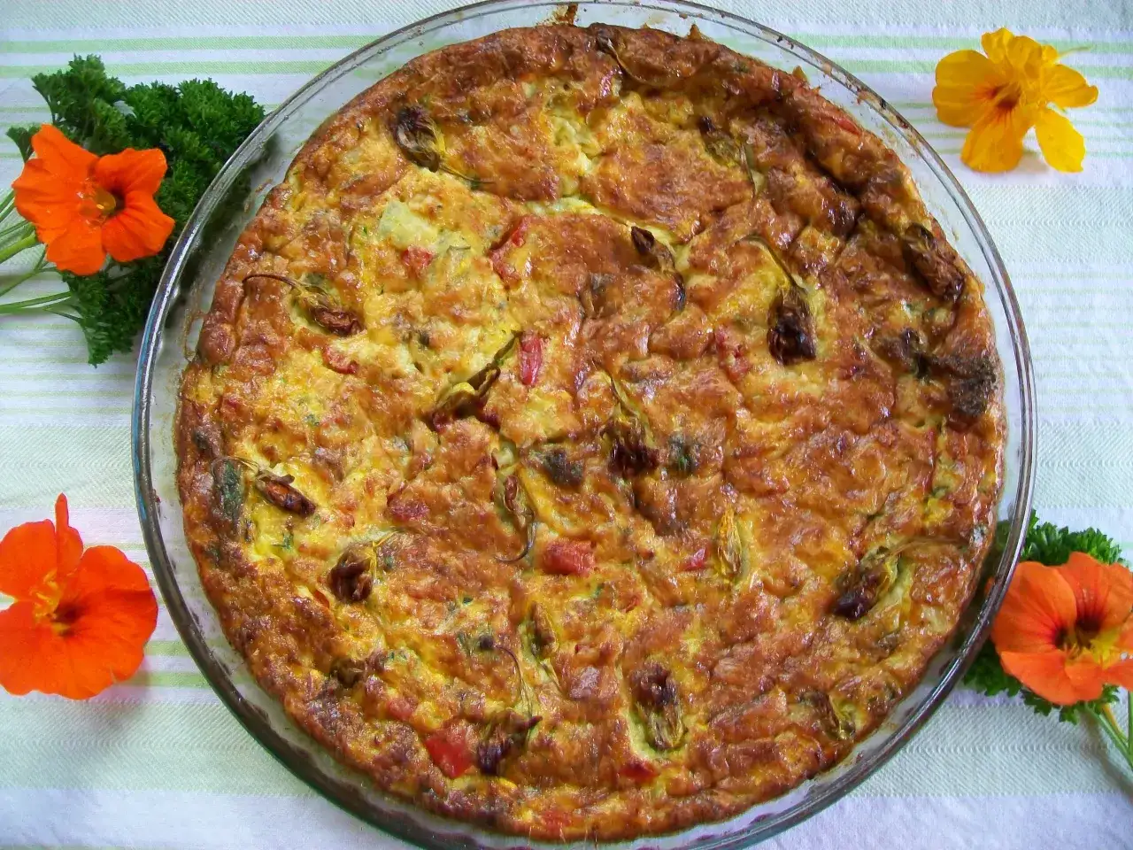 Złocista frittata z cukinią, pomidorami i ziołami w szklanej formie, ozdobiona nasturcjami i natką pietruszki.