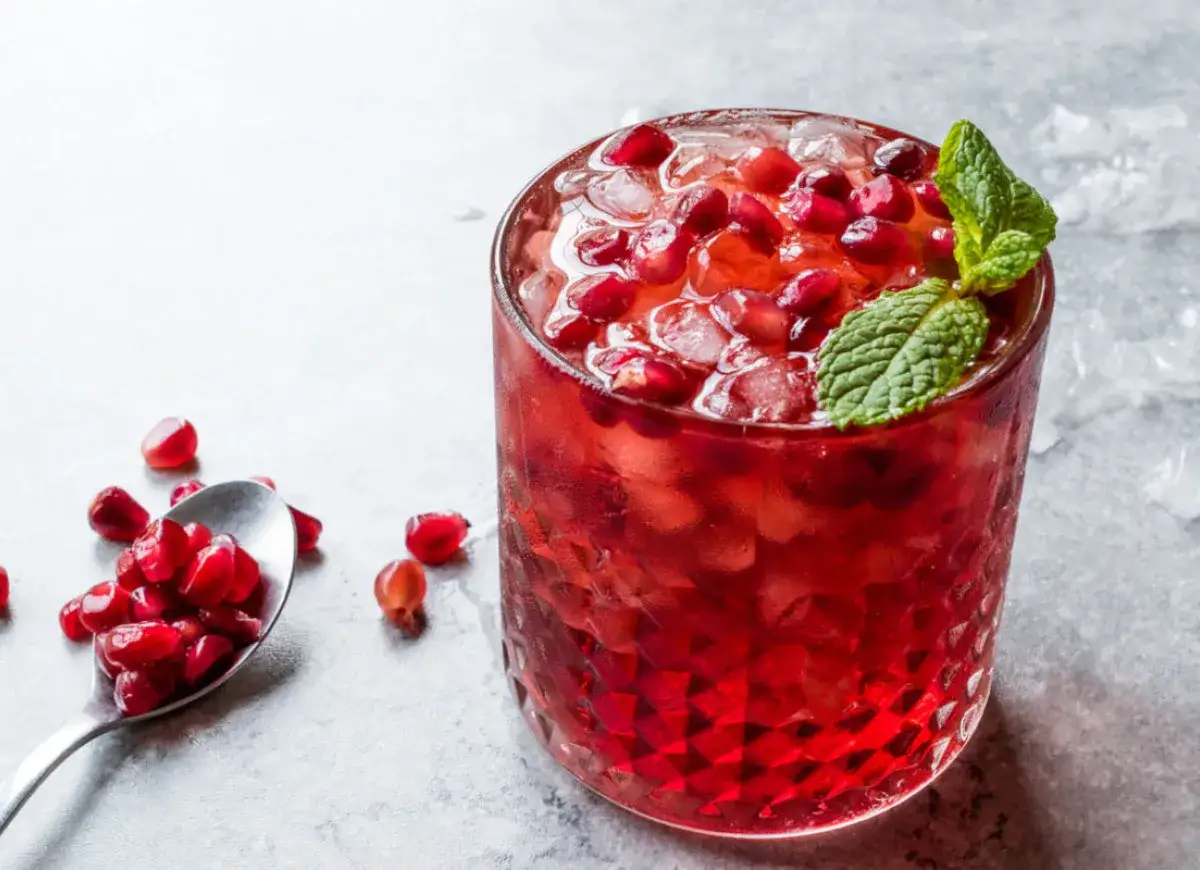 Orzeźwiający drink z grenadyną, lodem i pestkami granatu, ozdobiony listkiem mięty. Obok łyżeczka z owocem.