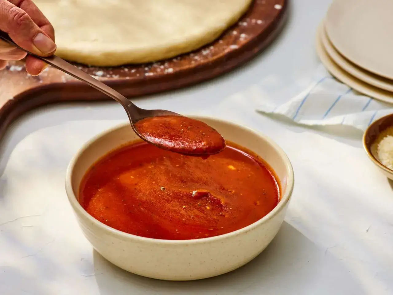 Łyżka nabiera gęsty, czerwony sos do pizzy z koncentratu. Obok leży ciasto na pizzę.