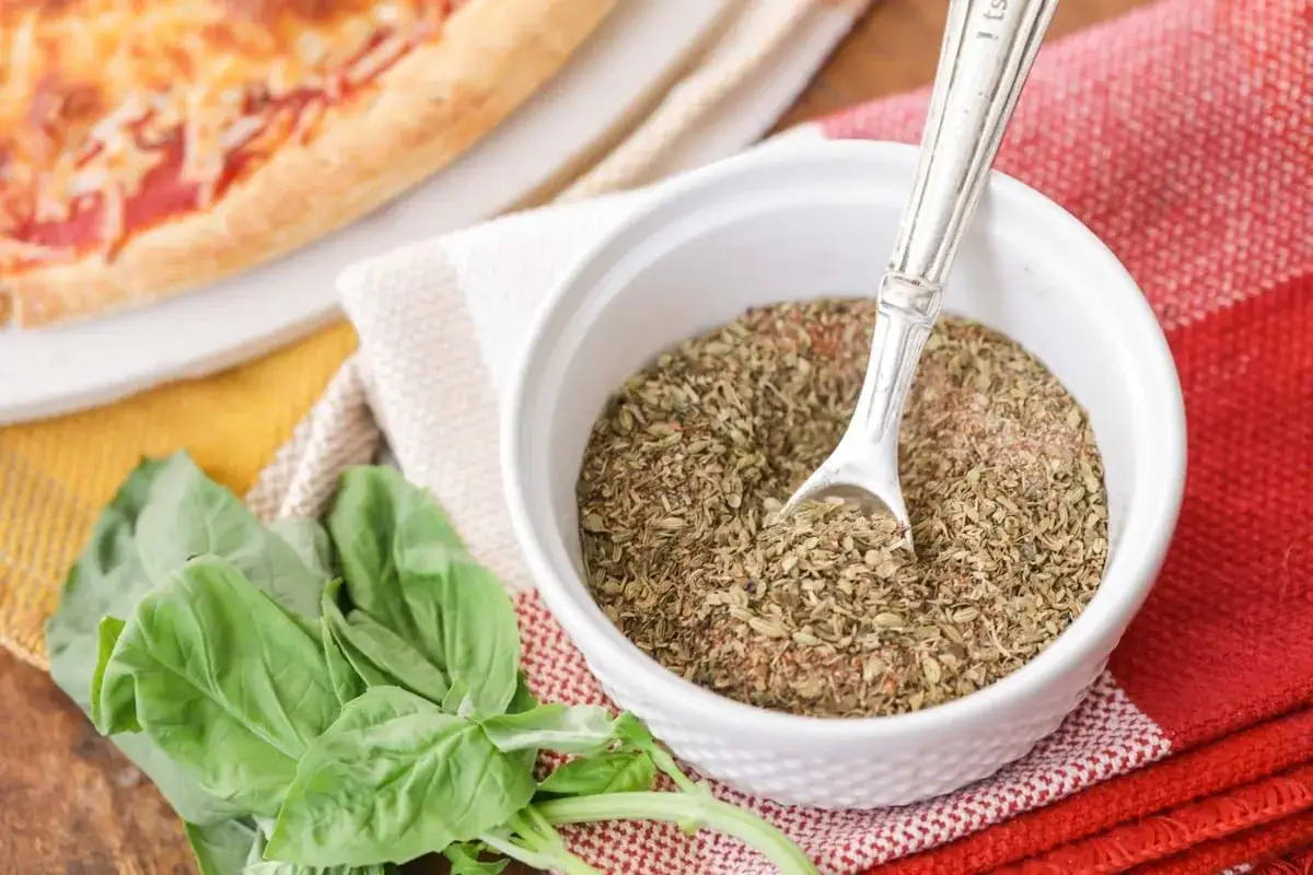 Mieszanka ziół, w tym oregano do pizzy, w białej miseczce z łyżeczką, obok świeżej bazylii i kawałka pizzy.