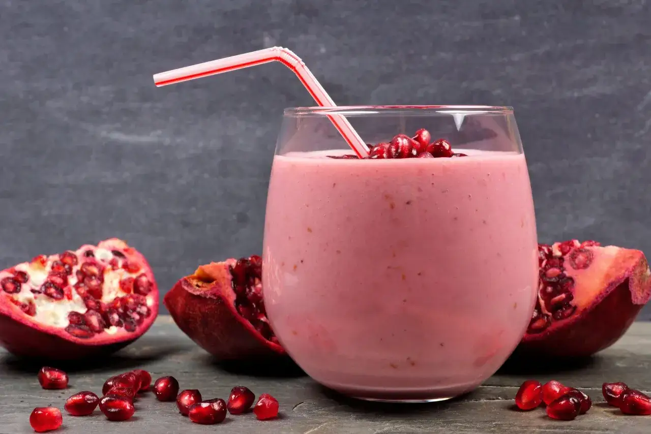 Różowy drink z granatem, ozdobiony pestkami, z czerwono-białą słomką, otoczony kawałkami granatu.