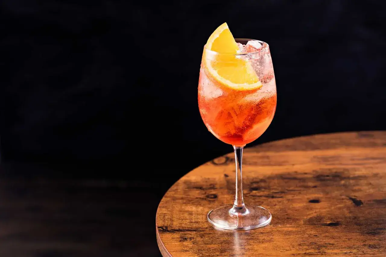 Orzeźwiający drink z Campari i Aperolem, ozdobiony plastrem pomarańczy, na drewnianym blacie.
