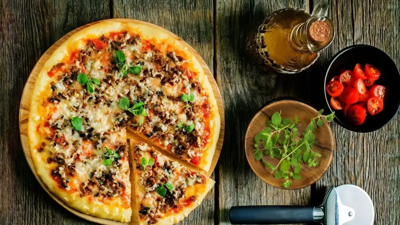 Pyszna pizza z mięsem mielonym, posypana świeżym oregano. Obok miseczka pomidorków i buteleczka oliwy. Idealna na wieczór z przyjaciółmi.