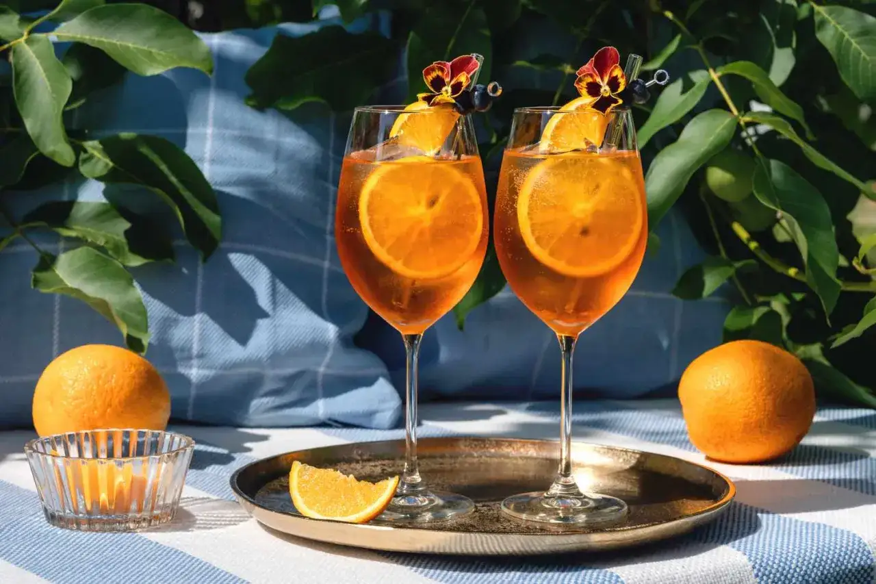 Dwa orzeźwiające drinki z aperolem i sokiem pomarańczowym, ozdobione plasterkami pomarańczy i kwiatami, gotowe do degustacji w letnim słońcu.