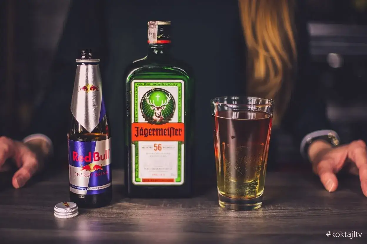 Jägermeister z czym pić? Klasycznie z Red Bullem, tworząc energetyczny koktajl. Na stole butelka Jägermeistera, Red Bull i szklanka z drinkiem.