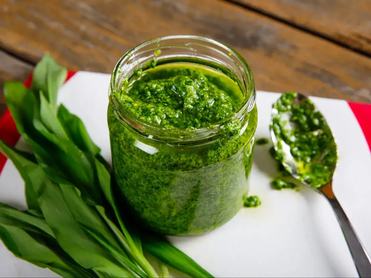 Słoik pełen aromatycznego pesto z czosnku niedźwiedziego, obok świeże liście i łyżka z zieloną pastą.