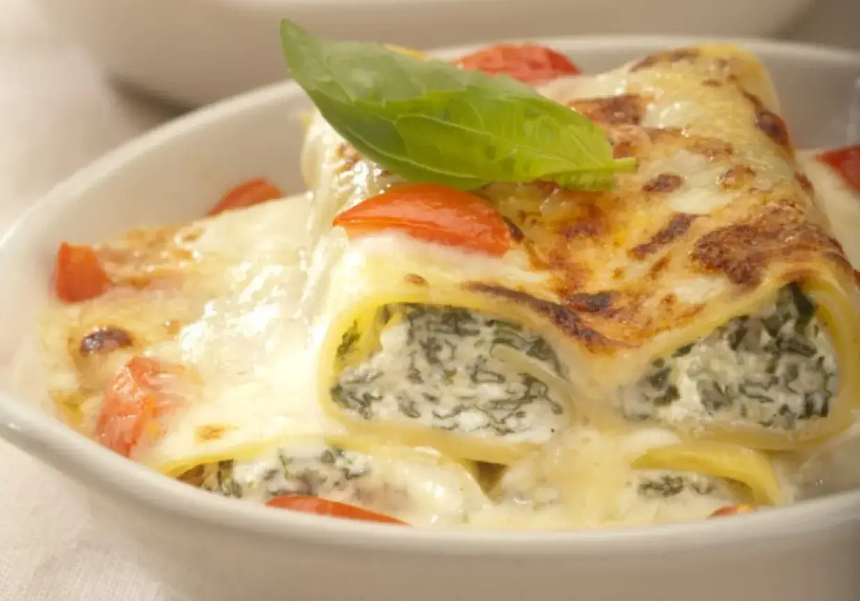 Pyszne cannelloni ze szpinakiem i serem, zapieczone pod beszamelem i pomidorami, ozdobione listkiem bazylii.