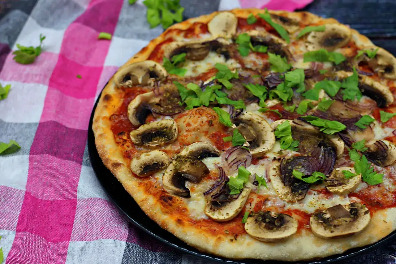 Pyszna pizza z pieczarkami i cebulą, posypana świeżą natką pietruszki, leży na czarnym talerzu, obok kraciastego obrusu.
