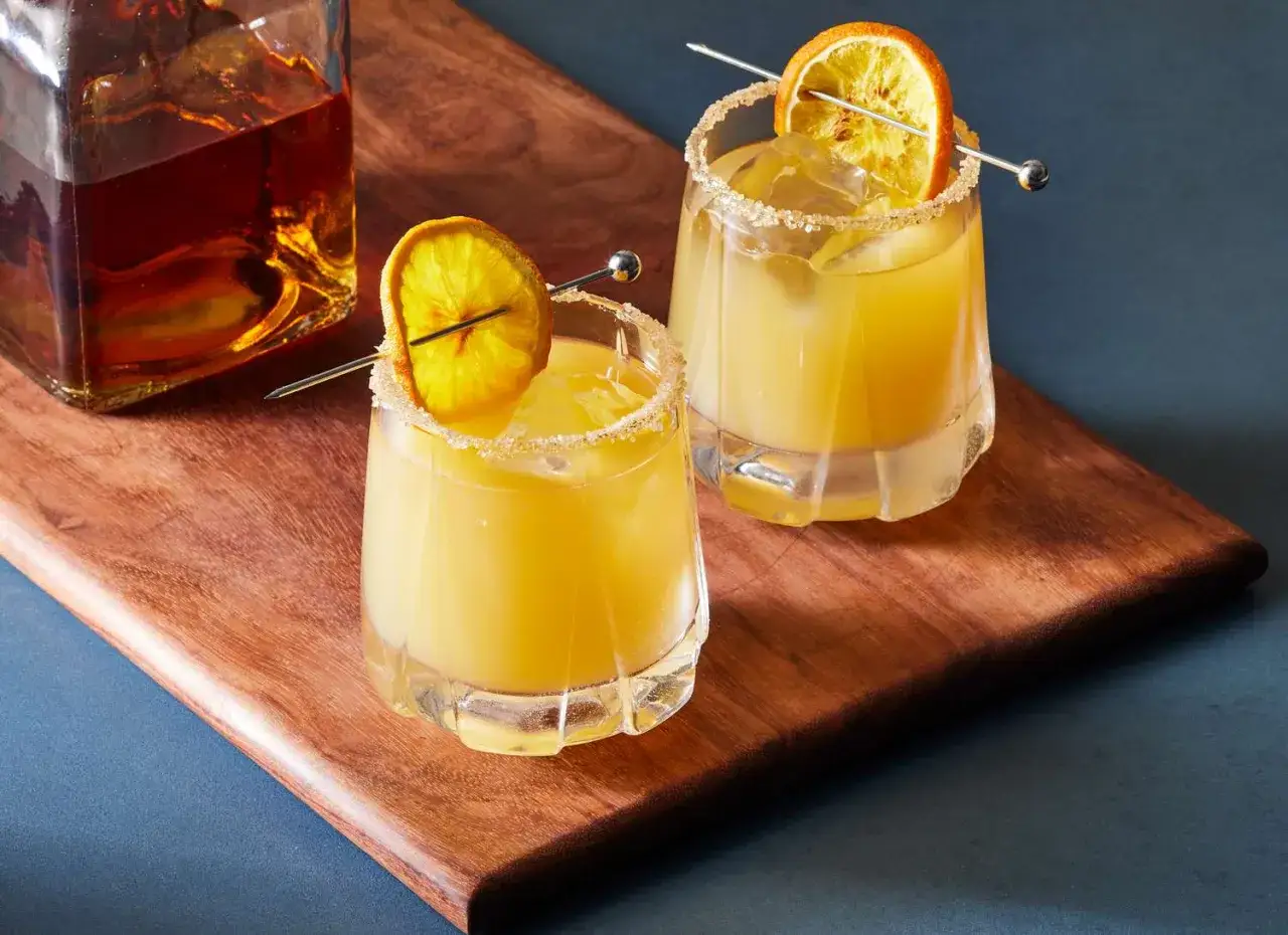 Dwa orzeźwiające whisky sour z plasterkiem pomarańczy i cukrowym rantem, obok butelki bursztynowego alkoholu.