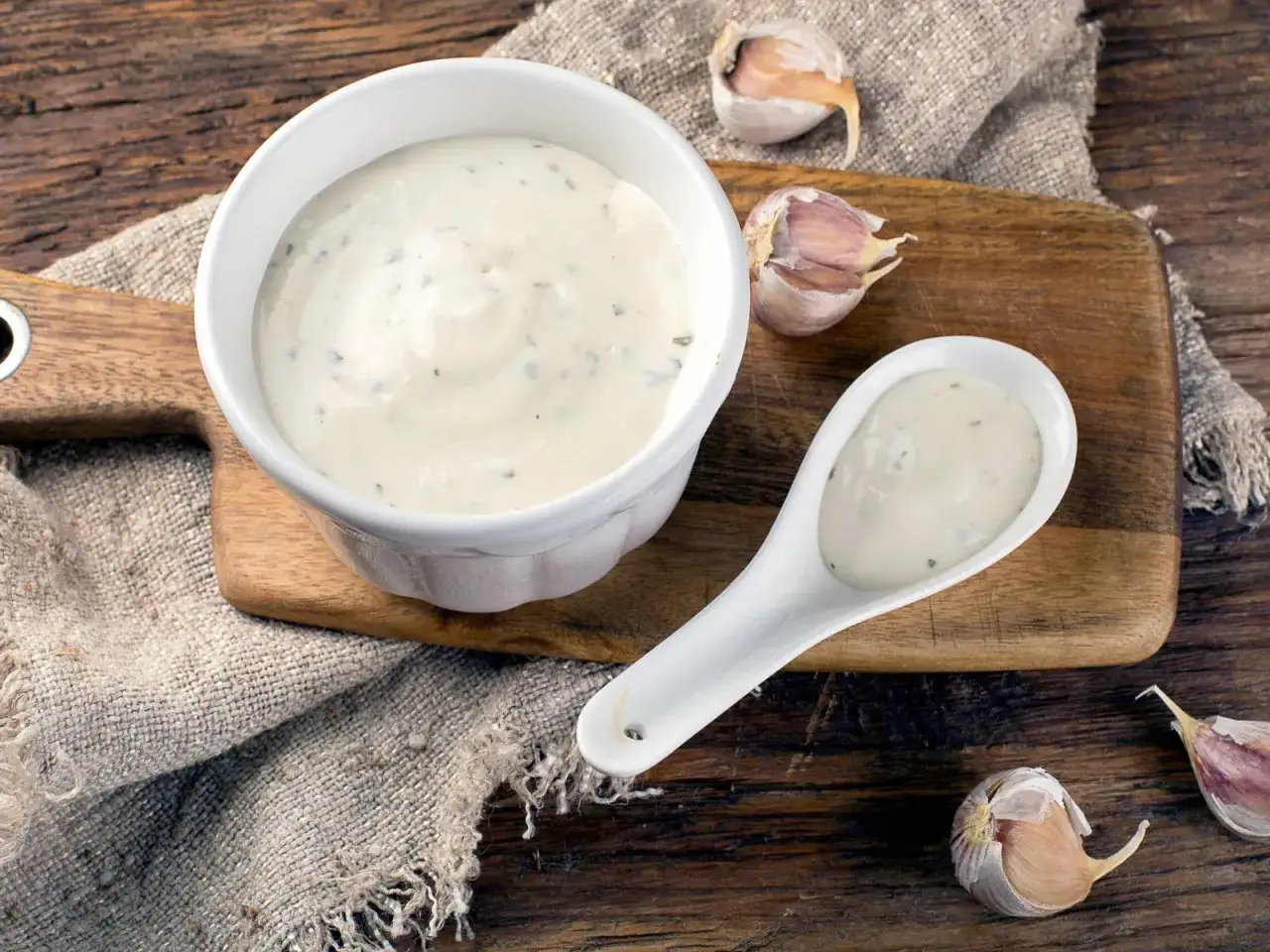 Kremowy sos aioli w miseczce i na łyżce, obok czosnku na drewnianej desce.