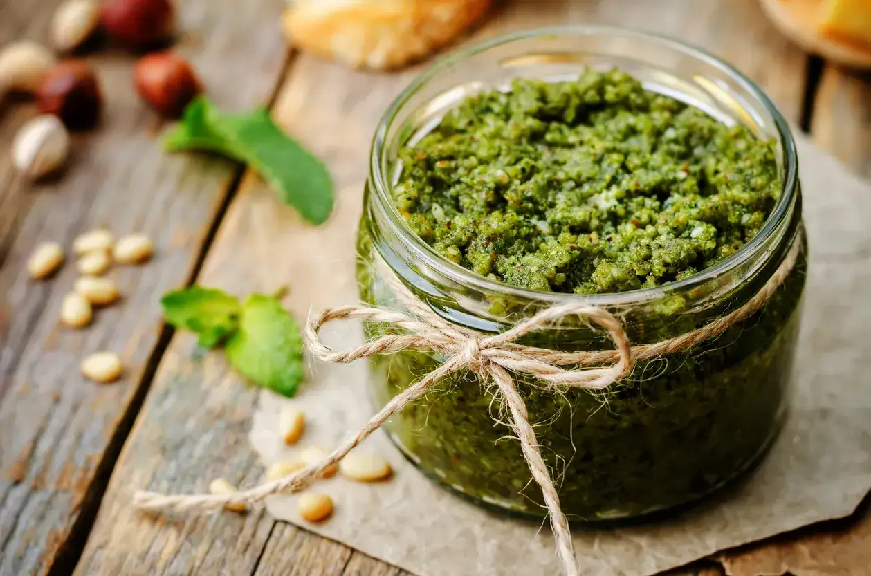 Słoik domowego pesto bazyliowego, ozdobiony sznurkiem, stoi na drewnianym stole. Wok&oacute;ł rozsypane są orzeszki piniowe i listki bazylii.