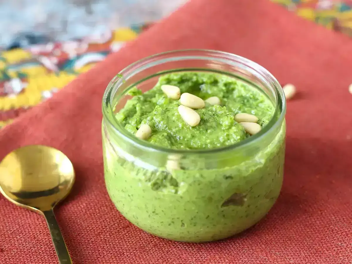 Słoiczek domowego pesto z rukoli, ozdobiony orzeszkami pinii. Obok złota łyżeczka.