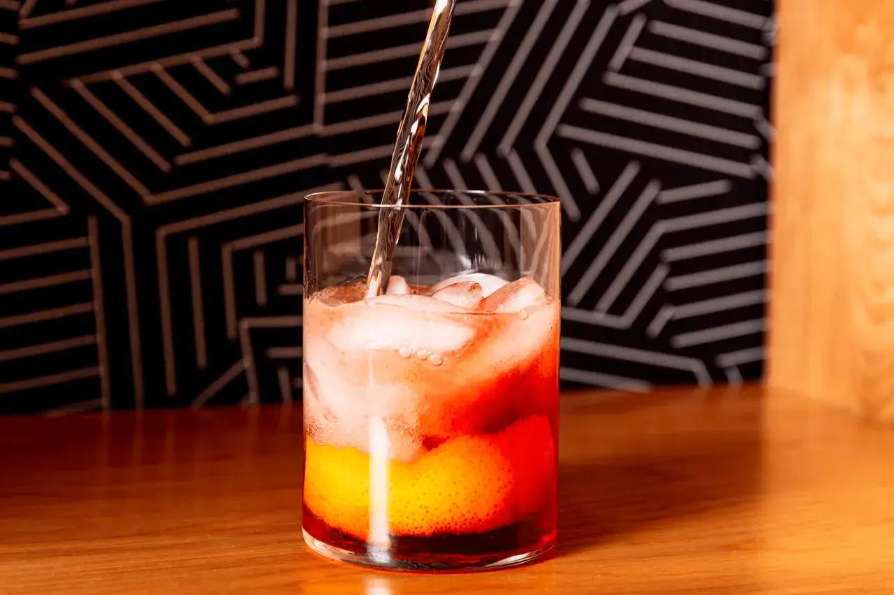 Przygotowanie Negroni Sbagliato: nalewanie prosecco do szklanki z lodem, pomarańczą i Campari.