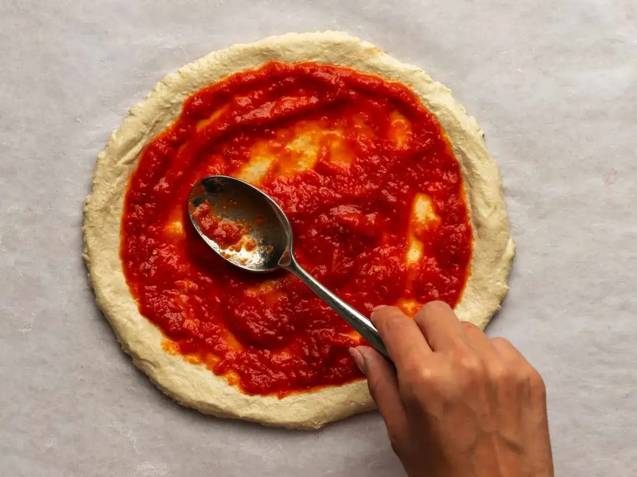 Ręka rozsmarowuje łyżką pyszny sos do pizzy z koncentratu na surowym cieście.