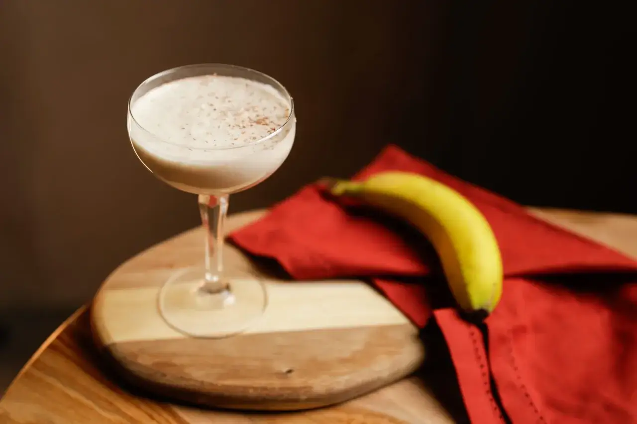 Kremowy likier bananowy drink, posypany cynamonem, obok dojrzałego banana na czerwonej serwetce.