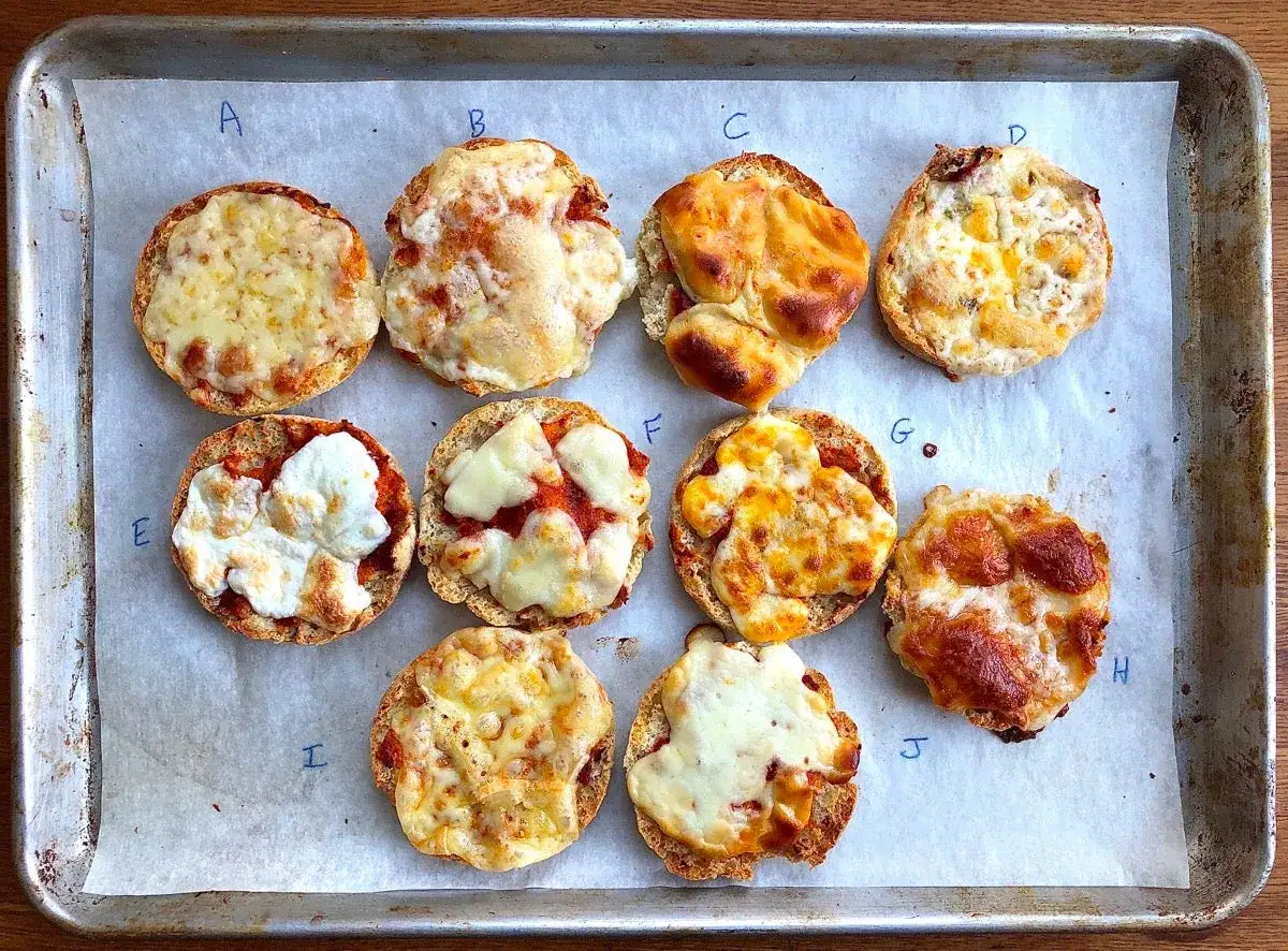 Mini pizze z różnymi rodzajami roztopionego sera na blasze. Każda z nich ma swój unikalny wygląd, niektóre z lekko przypieczonym serem, inne z bardziej kremowym.