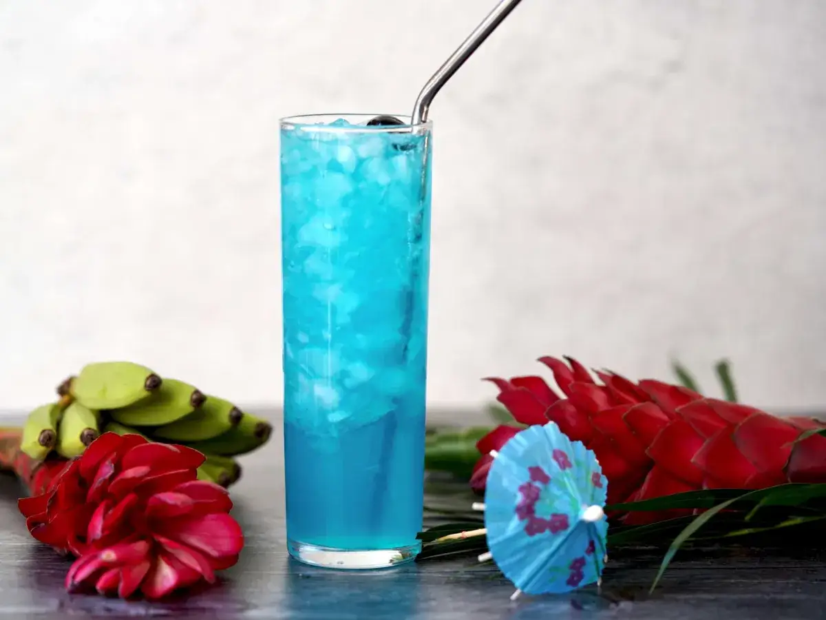 Letni drink z lodem i metalową słomką. Obok egzotyczne kwiaty i małe banany. Idealny przepis na blue curacao.