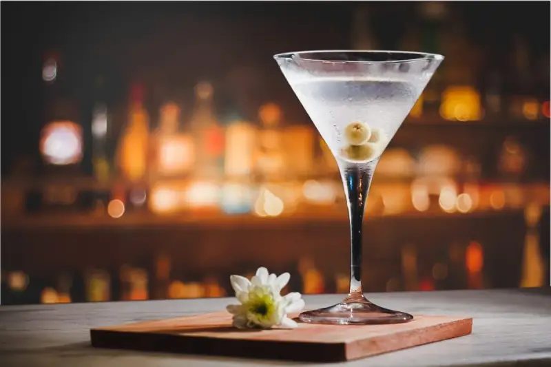 Eleganckie drinki z martini, ozdobione oliwkami, stoją na blacie baru. Obok kieliszka leży biały kwiat.